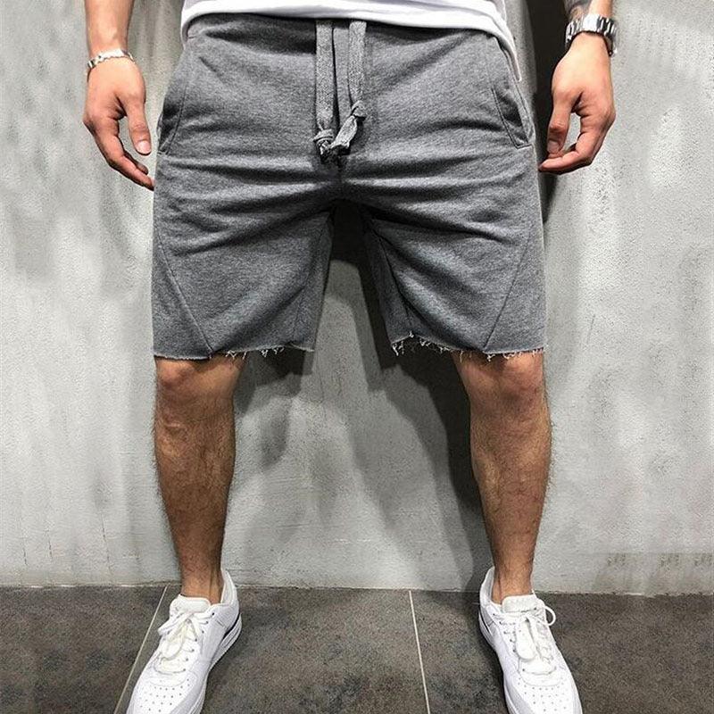 Man wearing casual grey drawstring shorts with raw hems and white sneakers against a gray wall