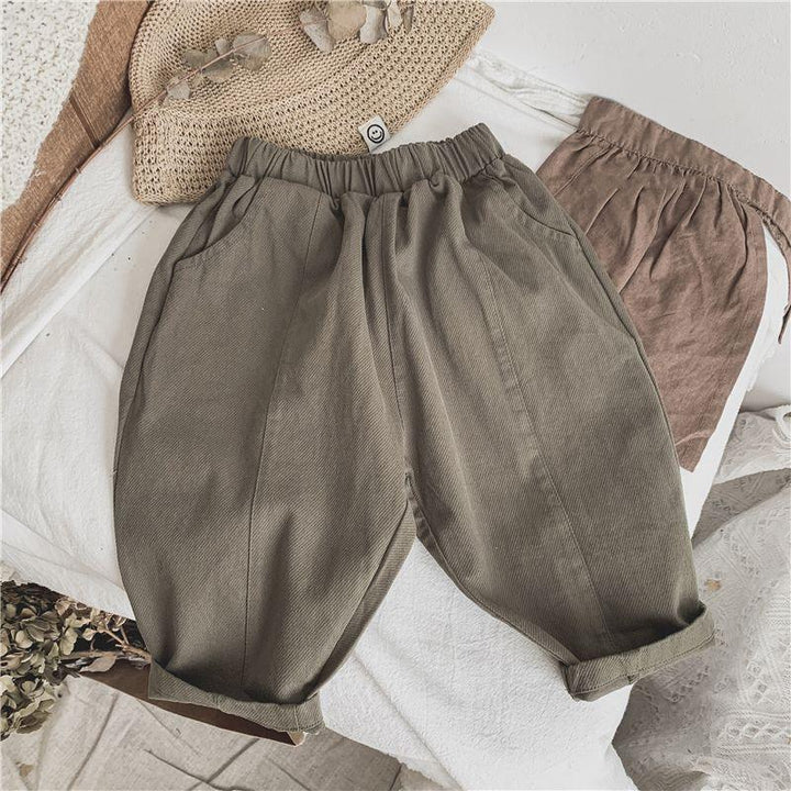 Casual olive green baby pants with elastic waistband on white textured fabric, straw hat and brown pouch nearby