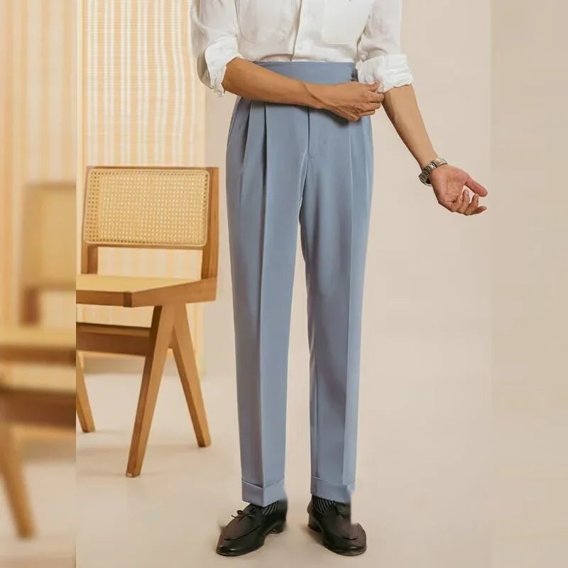 Man wearing light blue tailored trousers, white shirt, black dress shoes standing next to wooden chair