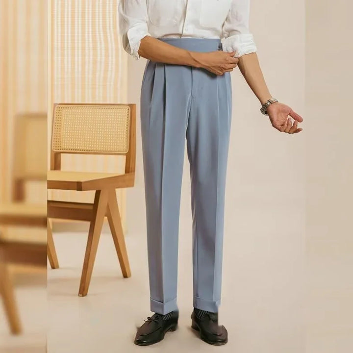 Man wearing light blue tailored trousers, white shirt, black dress shoes standing next to wooden chair