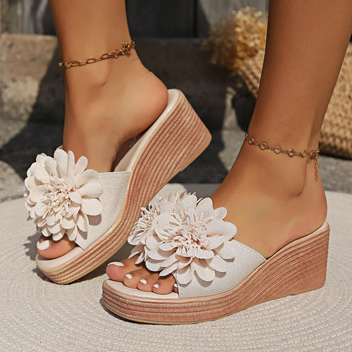 Wedge sandals with large white floral decoration, worn with gold ankle bracelets, outdoor setting