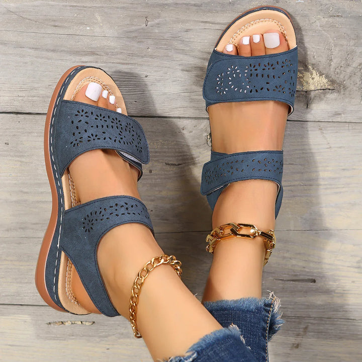 Blue open-toe sandals with laser-cut floral designs worn with gold chain anklets and frayed jeans on wooden floor