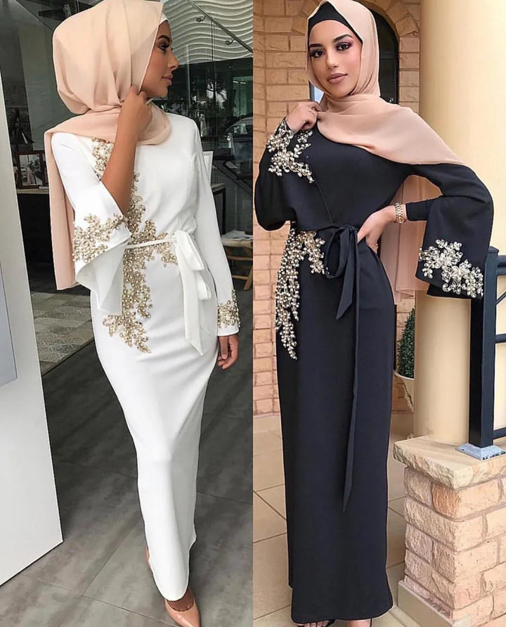 Two women wearing elegant modest dresses with gold embroidery and beige hijabs, one white and one black
