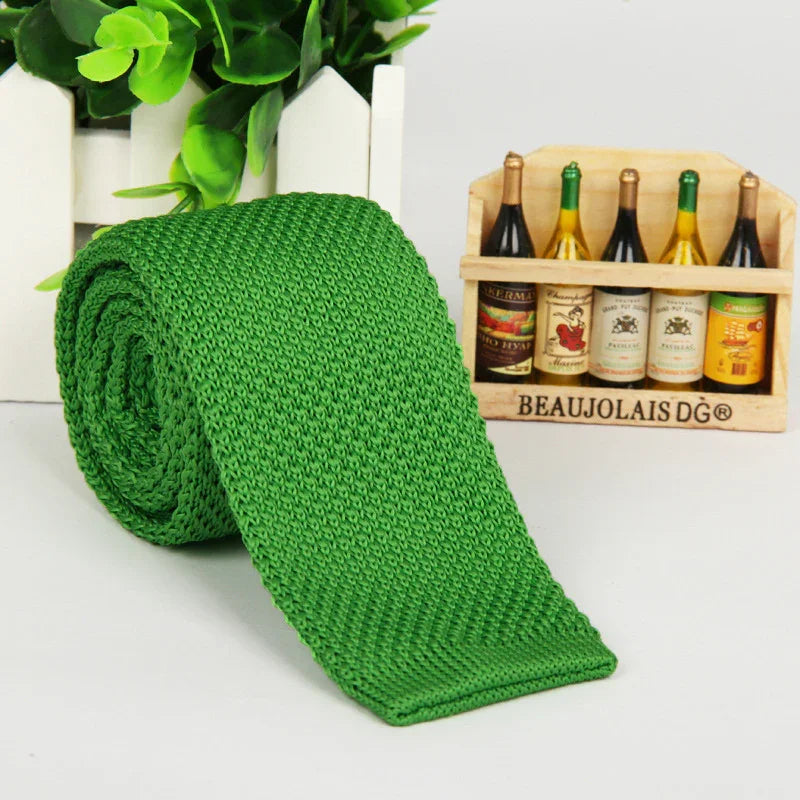 Green knitted tie rolled up on a white surface with a miniature wooden crate holding mini wine bottles and green leaves background