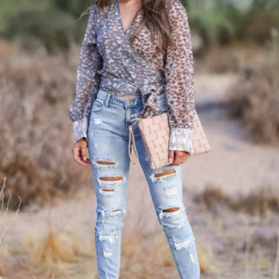 Woman wearing patterned wrap top with sheer sleeves, light blue ripped jeans, and holding a beige woven clutch outdoors