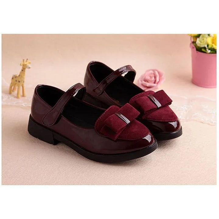 Burgundy patent Mary Jane shoes with velcro strap and suede bow detail on beige surface