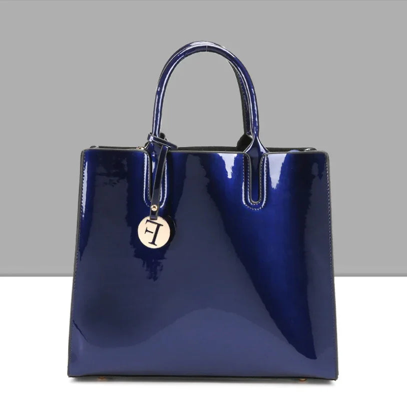 Glossy navy blue patent leather handbag with top handles and round gold charm on gray background