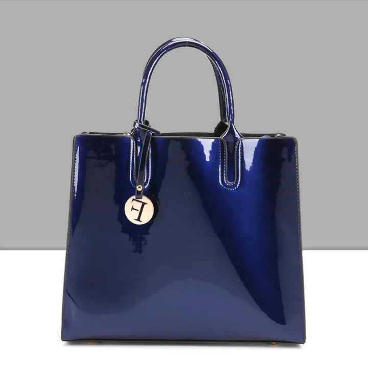 Glossy navy blue patent leather handbag with top handles and round gold charm on gray background