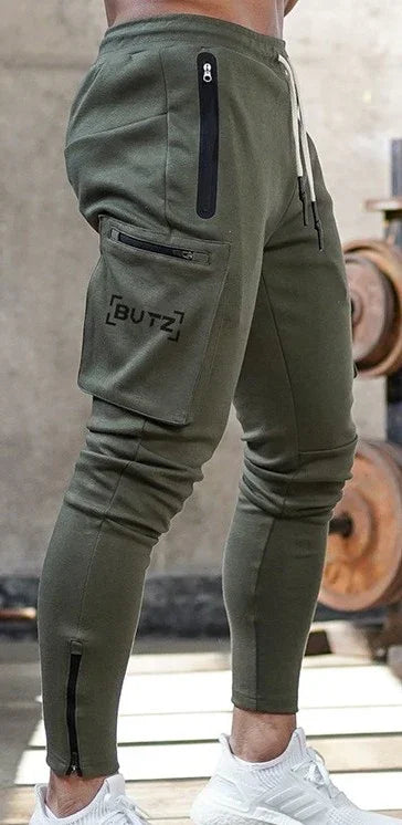 close-up of olive green men's jogger pants with zipper pockets and BUTZ logo, worn with white sneakers