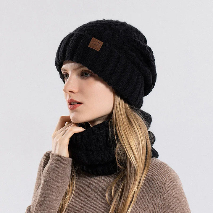 Woman wearing black knitted cable beanie and matching scarf with brown sweater on gray background