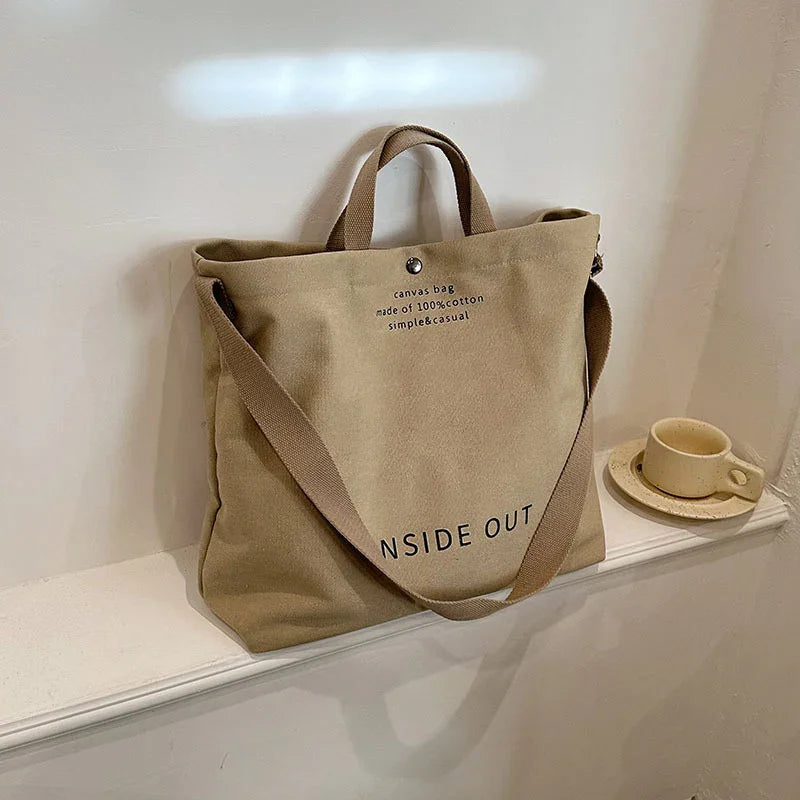 Beige 100% cotton canvas tote bag with handles and shoulder strap on white shelf next to ceramic cup and saucer