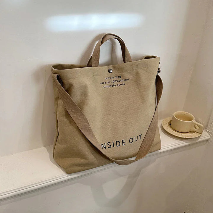 Beige 100% cotton canvas tote bag with handles and shoulder strap on white shelf next to ceramic cup and saucer