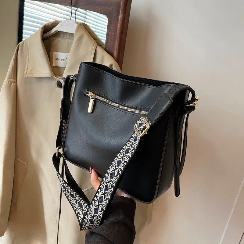 Black leather shoulder bag with gold zipper and patterned adjustable strap, beige jacket hanging nearby