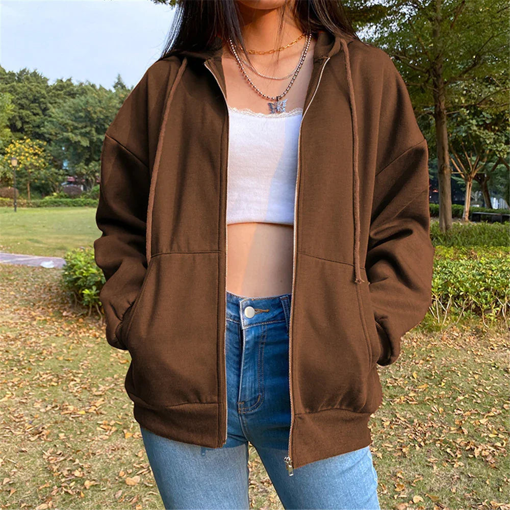 Woman wearing brown zip-up hoodie, white crop top, and blue jeans in outdoor park setting