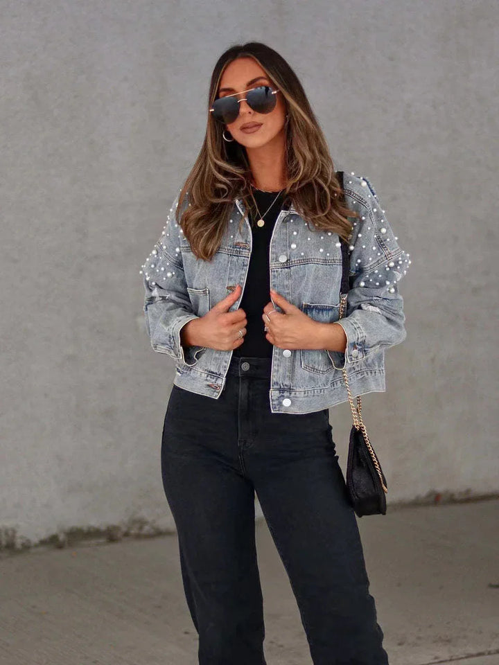 Woman wearing pearl embellished denim jacket, black top, black jeans, sunglasses, and shoulder bag against gray wall