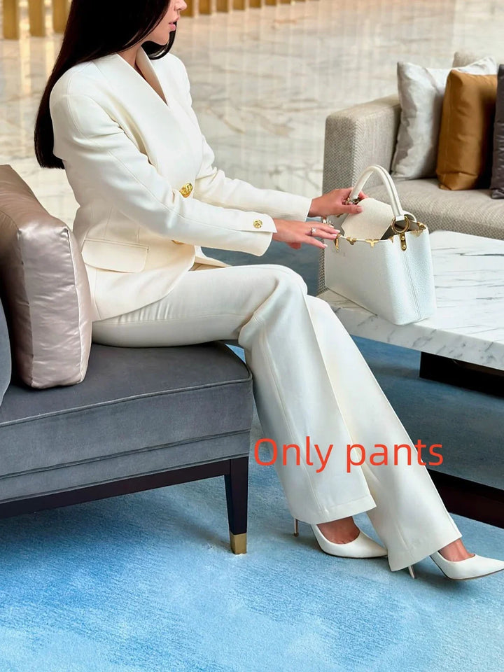 Woman in white tailored pants and blazer sitting on gray sofa reaching into white designer handbag