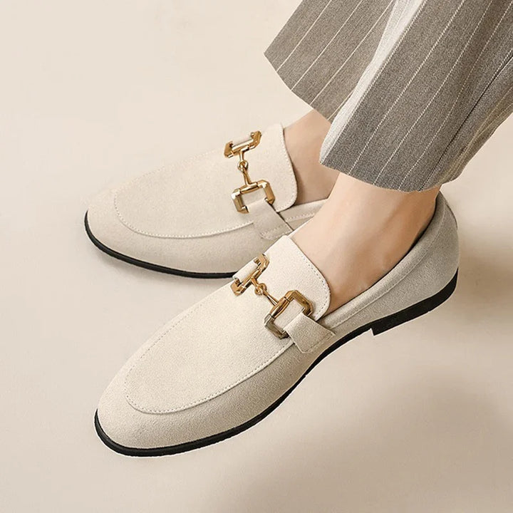 Beige suede loafers with gold horsebit detail worn with pinstripe trousers on a neutral background