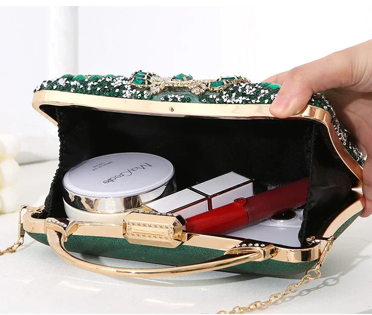Open green evening clutch bag with gold frame and bow detail, containing makeup items including compact and lipstick