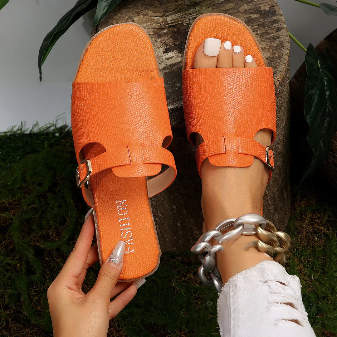 Orange textured slide sandals with buckle detail worn on foot with white nail polish and chunky bracelet