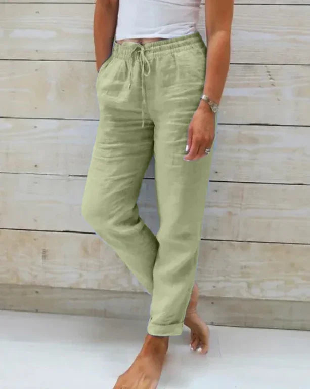 Woman wearing light green linen drawstring pants and white tank top against wood wall