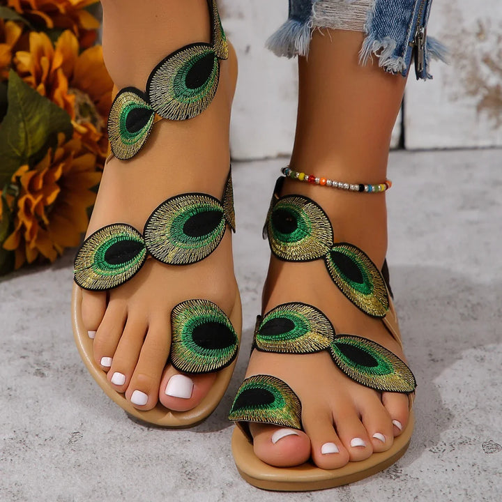 Close-up of feet wearing studded sandals with green and gold peacock feather designs and white nail polish