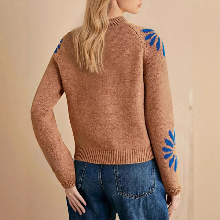 Woman wearing brown knitted sweater with blue flower designs on sleeves, paired with blue jeans