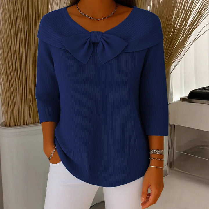 Woman wearing navy blue sweater with large bow detail and 3/4 sleeves paired with white pants
