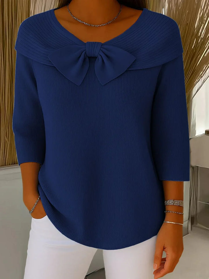 Woman wearing navy blue ribbed sweater with large bow detail and white pants indoors
