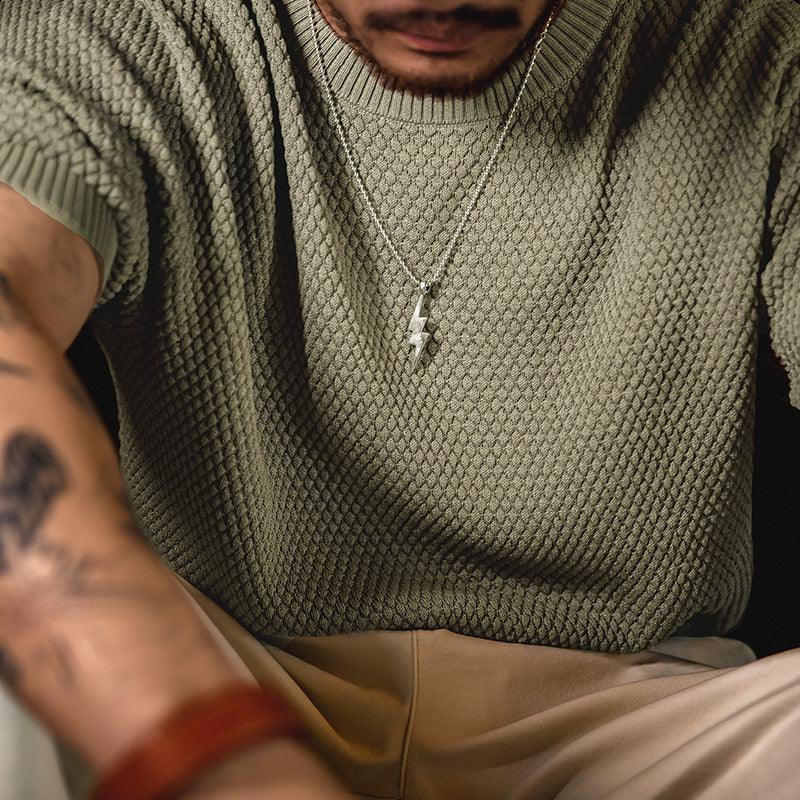 Close-up of man wearing olive textured knit sweater, lightning bolt pendant necklace, and beige pants