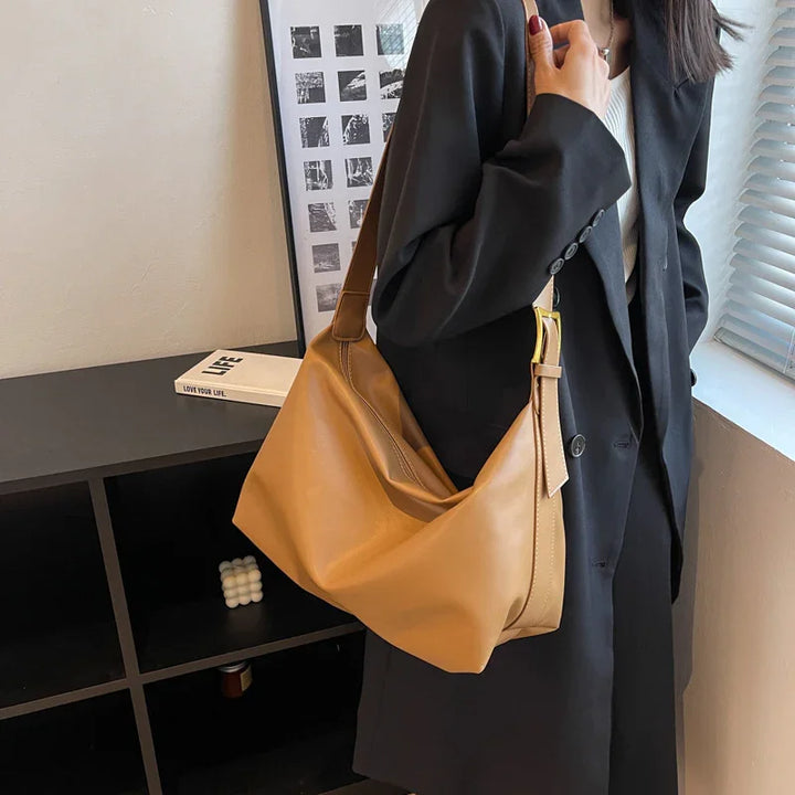 Woman in black coat holding a tan leather shoulder bag indoors near window and shelf