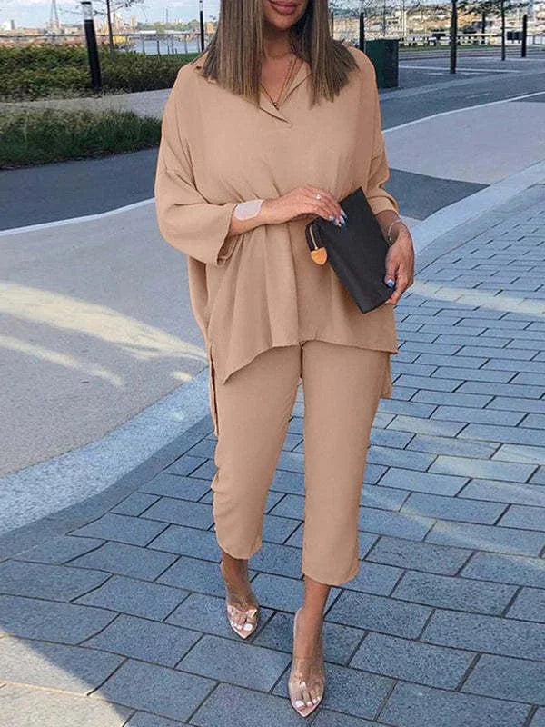Woman wearing beige two-piece outfit with loose top and cropped pants, clear heels, holding black clutch outdoors
