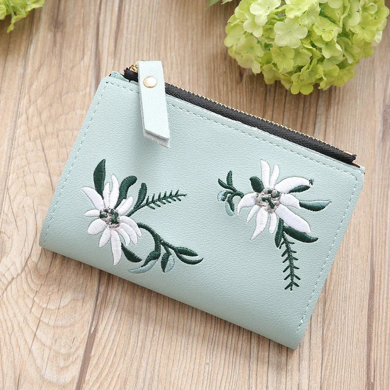 Light blue leather wallet with white floral embroidery on wooden surface with green flowers