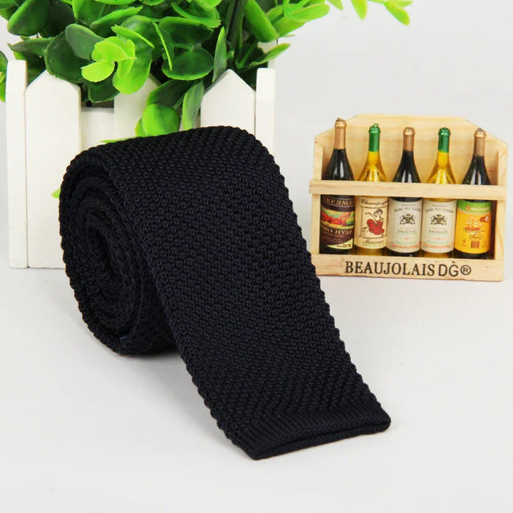 Black knitted men's tie rolled up next to a wooden wine bottle holder with miniature bottles