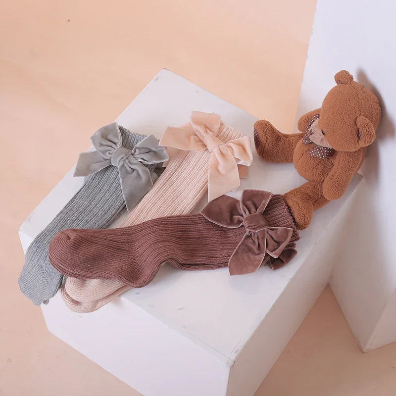 Three ribbed knit socks in gray, beige, and brown with large velvet bows, next to a brown teddy bear