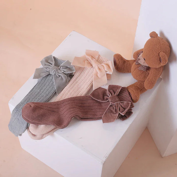 Three ribbed knit socks in gray, beige, and brown with large velvet bows, next to a brown teddy bear