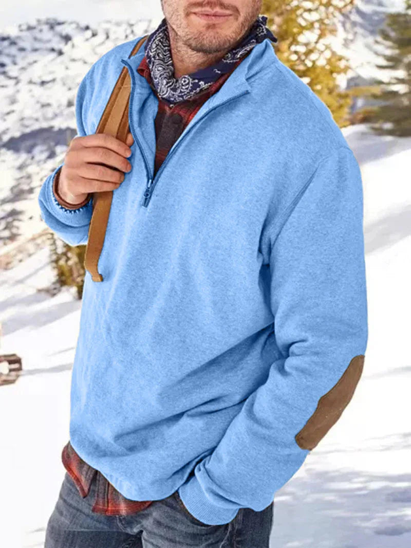 Man wearing light blue quarter-zip fleece sweater with elbow patches and bandana in snowy mountain setting