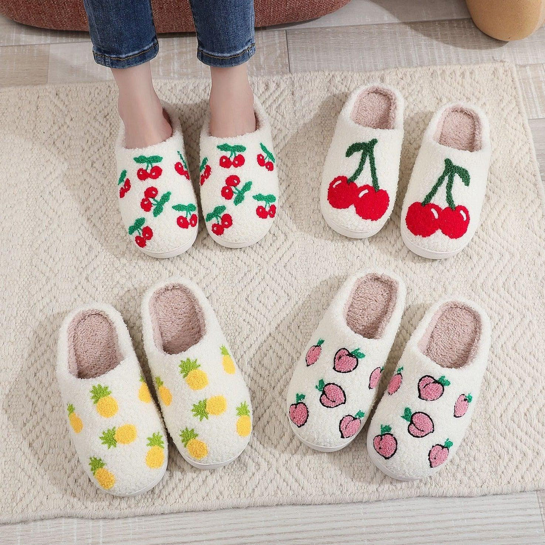 Four pairs of white cozy slippers with fruit designs: cherries, pineapples, and peaches on a textured rug