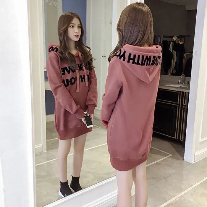 Young woman wearing a dusty pink oversized hoodie dress with black text, standing in front of a full-length mirror indoors