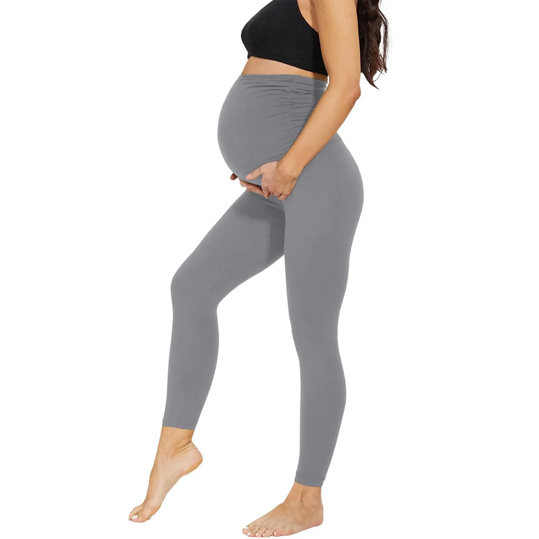 Pregnant woman wearing gray maternity leggings and black sports bra standing barefoot on white background