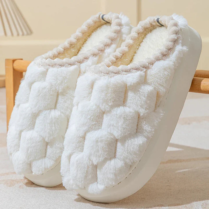 Pair of white plush indoor slippers with textured soft fur and cushioned soles on a beige surface