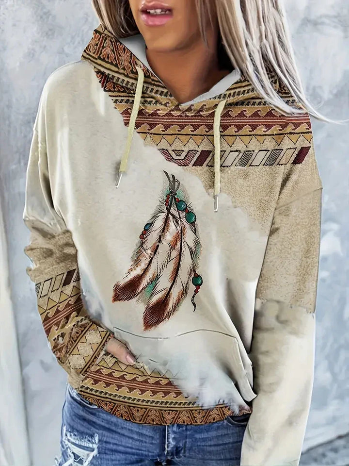 Woman wearing beige hoodie with tribal patterns and print of feathers with turquoise beads, paired with distressed blue jeans