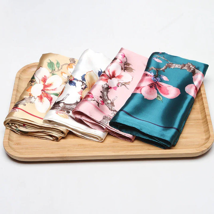 Four floral patterned silk scarves in beige, white, pink, and teal folded on a wooden tray