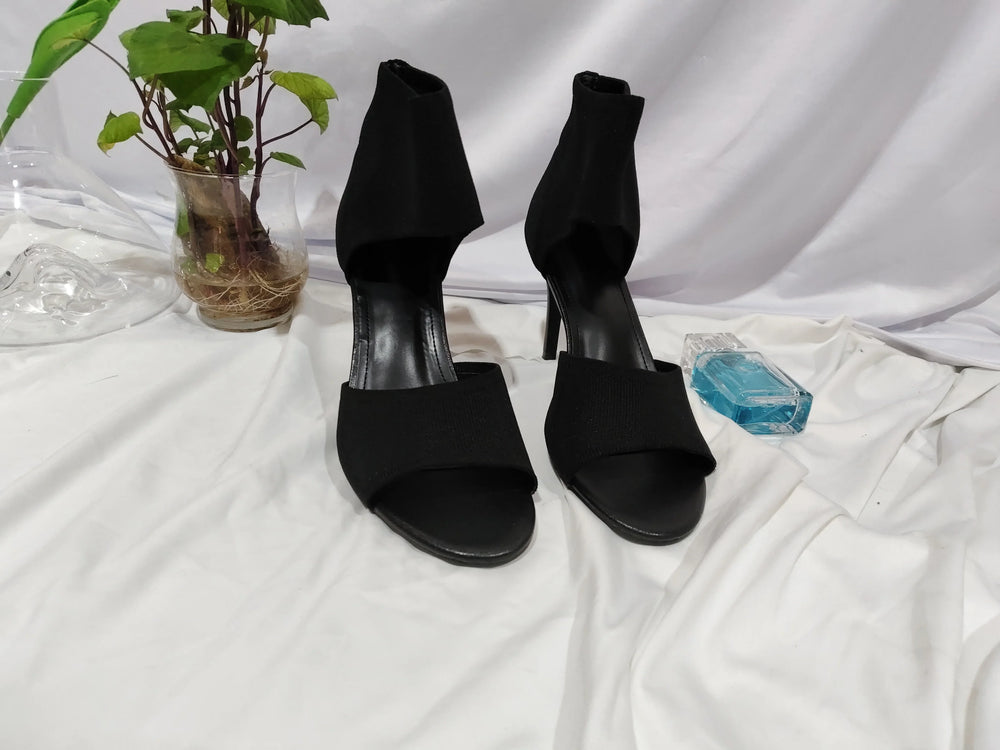 Pair of black high-heeled sandals on white cloth with green plant in glass vase and blue perfume bottle
