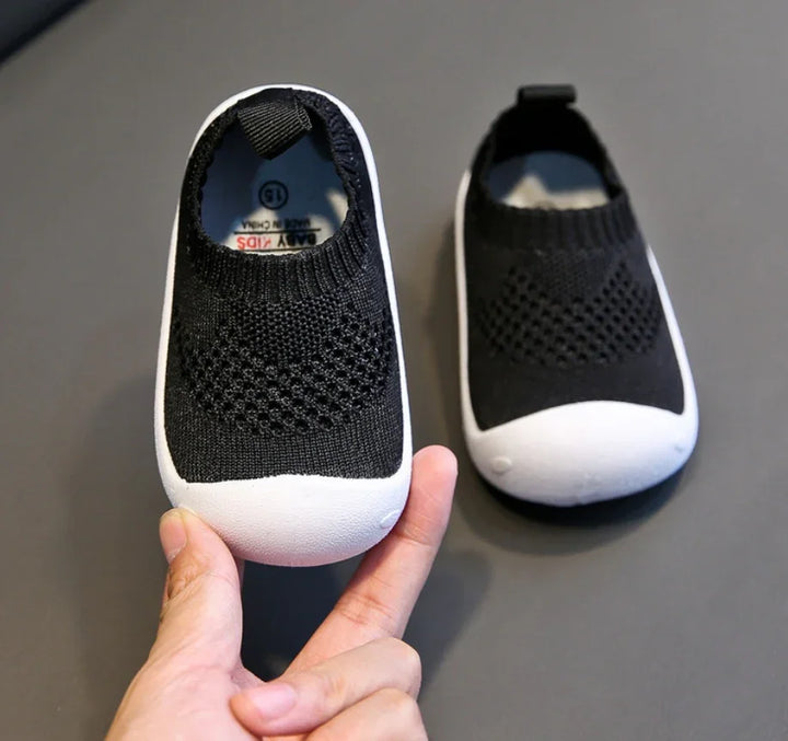 Hand holding black baby shoes with white soles on grey background