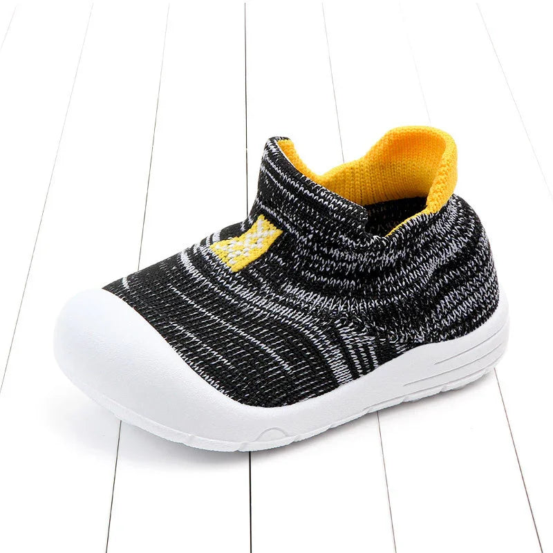 Black and white knit toddler shoe with yellow inner lining and white rubber sole on white wooden floor