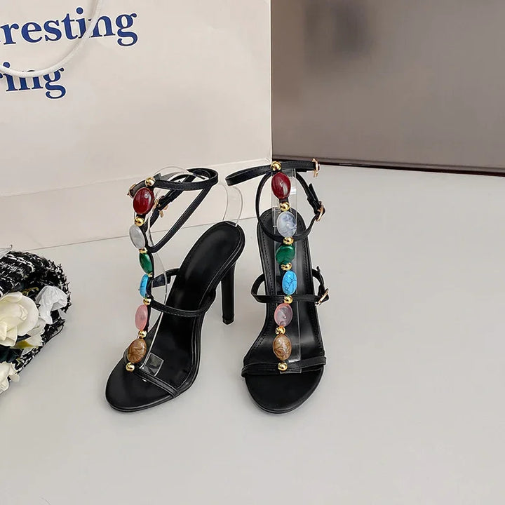Black high-heeled sandals with multicolored gemstone embellishments and ankle straps on white surface