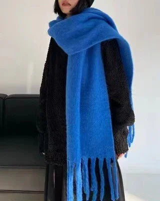 Person wearing a large blue knit scarf with fringe over a black textured sweater indoors