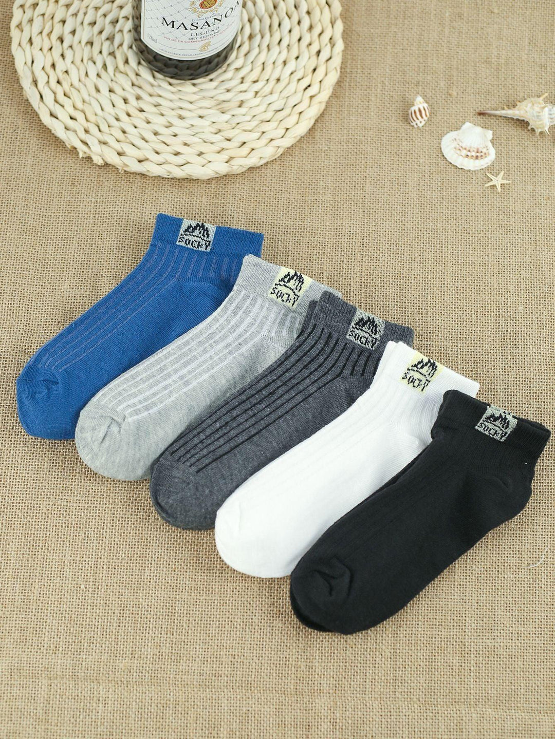 Set of five ankle socks in blue, gray, dark gray, white, and black on burlap fabric with seashells and woven mat
