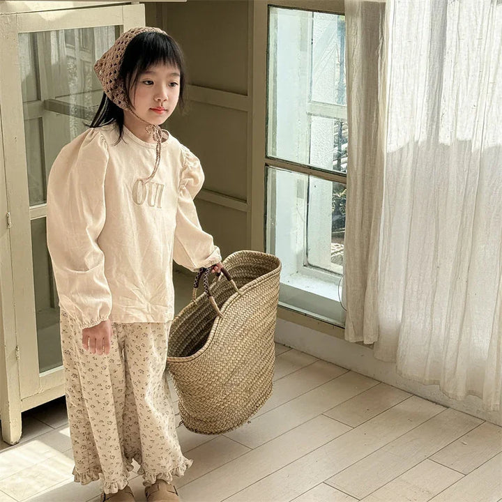 Young girl in beige puff-sleeve top and floral pants holding woven basket in sunlit room