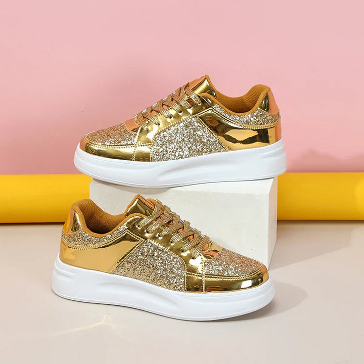 Pair of gold glitter and metallic sneakers with white soles on white block, pink and yellow background